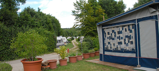 Eine Reihe von Topfpflanzen neben einem Wohnwagen mit weiteren Wohnwagen entlang eines Kiesweges, umgeben von Bäumen und Grünpflanzen.