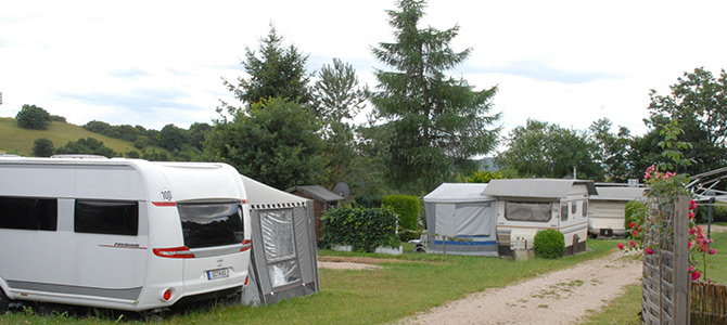 Ein kleiner Wohnwagenpark mit mehreren Wohnwagen und Zelten, die auf grasbewachsenen Parzellen aufgestellt sind, die von Bäumen und einem Kiesweg begrenzt werden, unter einem bewölkten Himmel.