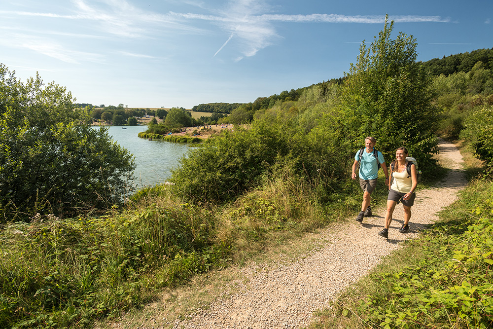 Zwei Personen gehen auf einem Kiesweg neben einem See, umgeben von Grün und Bäumen unter einem klaren, blauen Himmel.