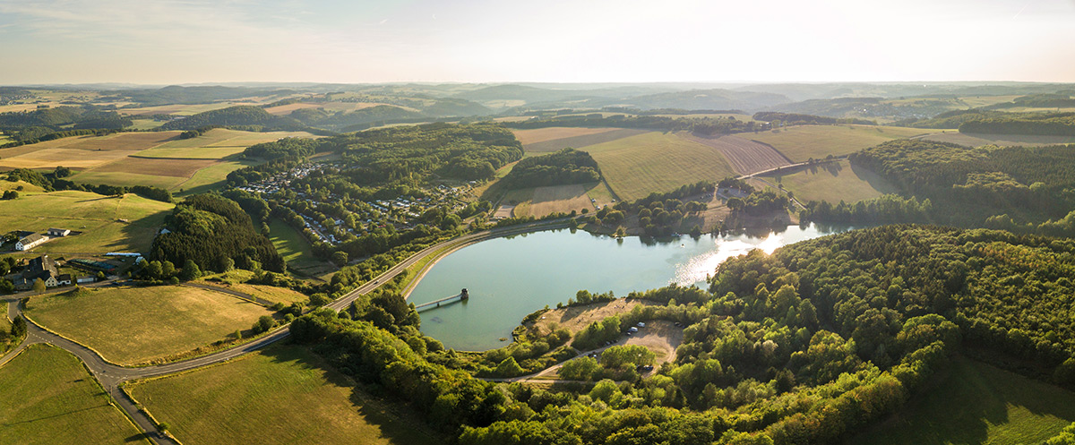 Luftaufnahme einer ländlichen Landschaft mit einem großen spiegelnden See, umgeben von Feldern, Wäldern und verstreuten Gebäuden unter einem klaren Himmel.
