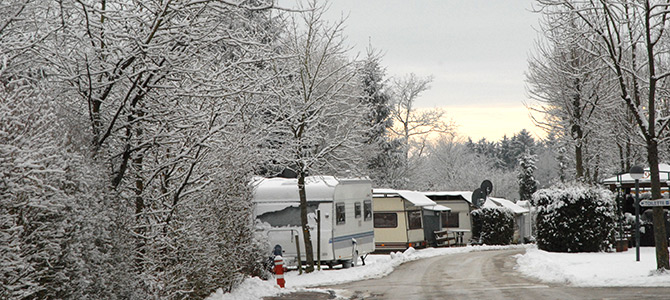 Schneebedeckte Wohnmobile und Bäume säumen eine kurvenreiche Straße auf einem Wintercampingplatz, der Himmel ist bedeckt und auf dem Boden liegen Schneeflecken.