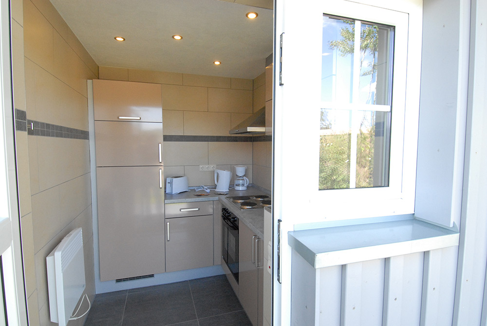 Kleine moderne Küche mit beigen Schränken, einem Herd, einem Kühlschrank, einer Mikrowelle und einer Kaffeemaschine, Blick durch ein offenes Fenster.