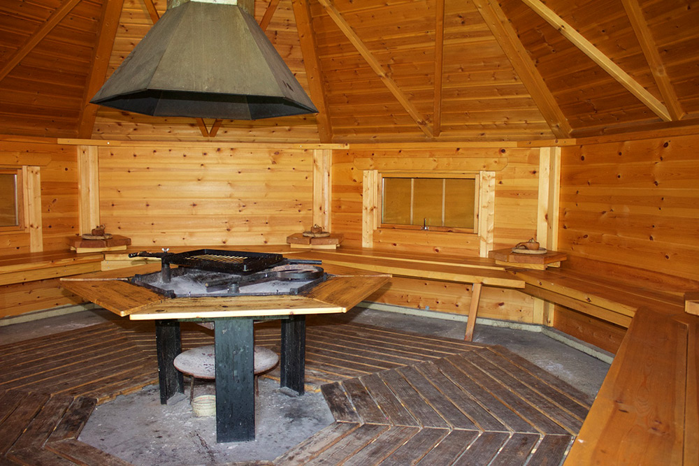 Ein achteckiger Raum aus Holz mit eingebauten Bänken, einem zentralen Grill und Kamin sowie kleinen Fenstern an den Wänden.