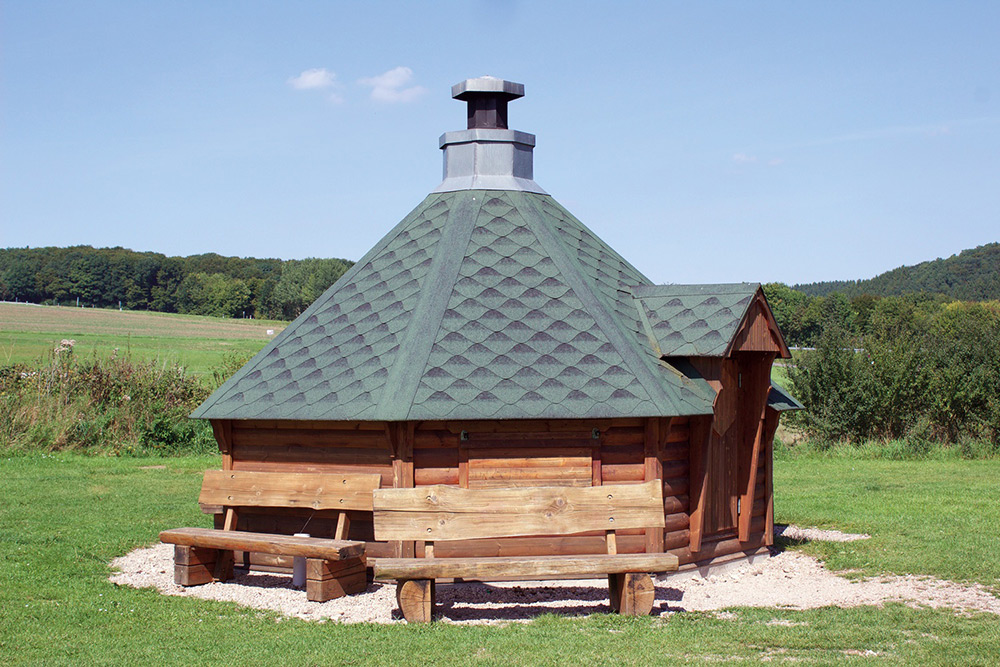 Eine sechseckige Holzhütte mit grünem Schindeldach und Schornstein steht auf einer Wiese mit zwei Bänken davor und Bäumen im Hintergrund.