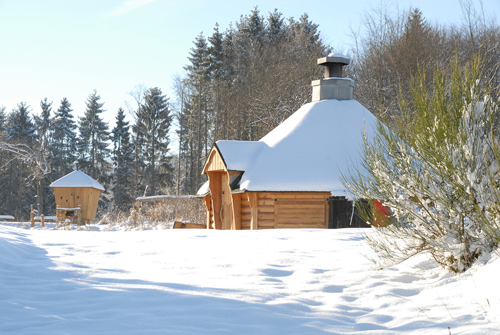 Eine Holzhütte mit Schornstein steht in einer verschneiten Landschaft, umgeben von Bäumen und unter einem klaren Himmel.