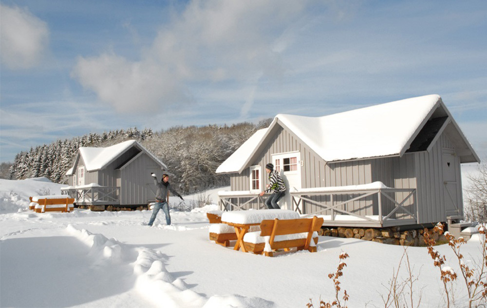 Zwei Personen liefern sich eine Schneeballschlacht vor zwei kleinen Hütten, umgeben von schneebedeckten Bänken, Bäumen und einer Landschaft unter blauem Himmel.