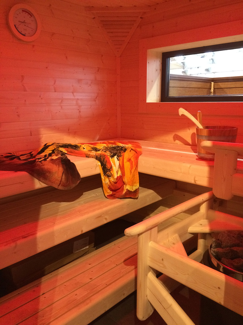 Eine Holzsauna mit Bänken, einem bunten Handtuch, einem Eimer mit einer Kelle, einem kleinen Fenster und einem runden Thermometer an der Wand.