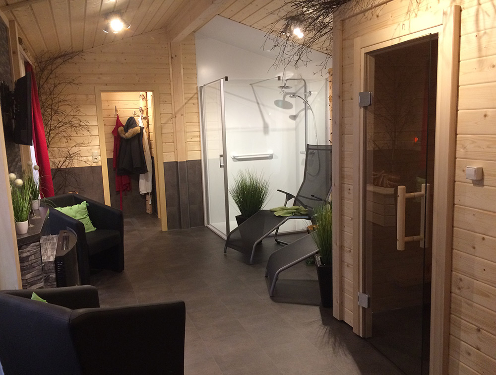 Moderner Wellness-Raum mit Holzwänden, Liegestühlen, einer verglasten Dusche, einer Sauna, einer Sitzecke mit Kissen und Mänteln, die am Eingang hängen.