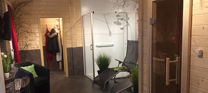 A modern spa room with wooden walls, two lounge chairs, a glass shower enclosure, and direct access to a sauna, with coats hanging in the hallway.