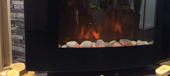 A modern electric fireplace with a black frame, white stones at the base, and a simulated flame effect, set against a stone-patterned wall with cut wood stacked nearby—ideal for creating a cozy sauna-inspired retreat.