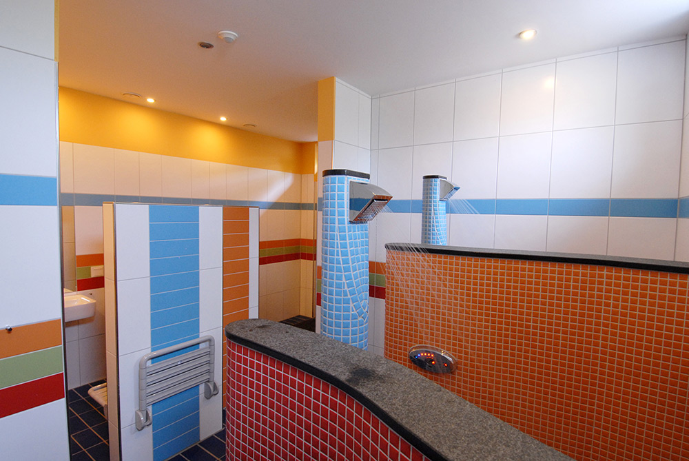 Öffentlicher Duschbereich mit gefliesten Wänden, zwei Duschköpfen, Sichtschutzwänden und einem Farbkonzept in Blau, Orange und Weiß.