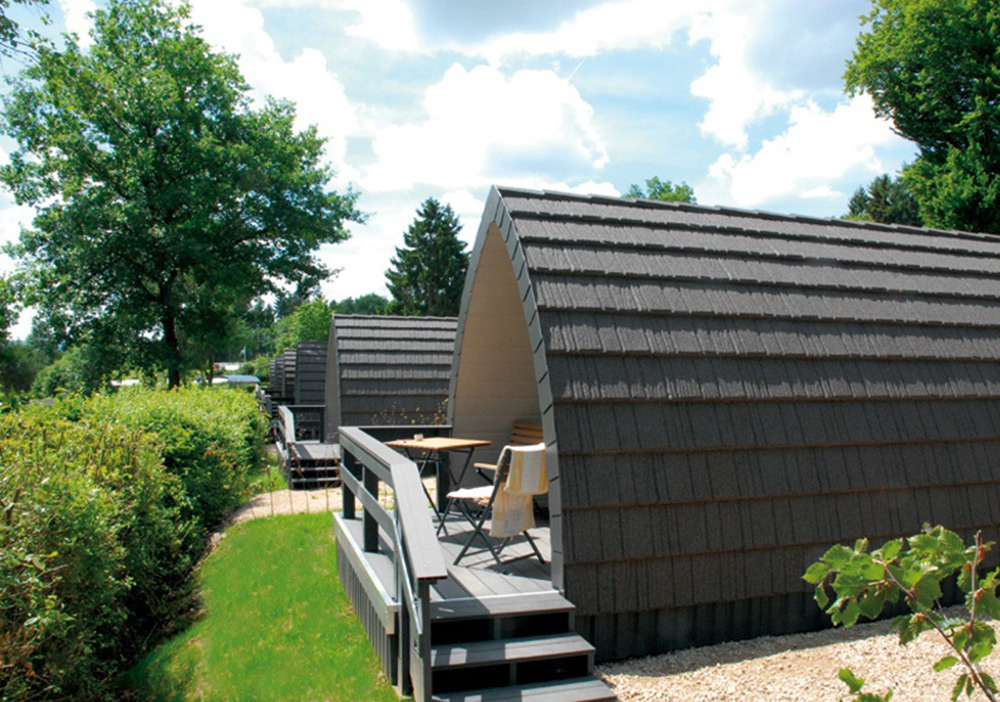 Kleine, moderne Hütten mit geschwungenen Dächern sind aneinandergereiht, jede mit einer Veranda und Sitzgelegenheiten im Freien, umgeben von Grün und Bäumen unter einem teilweise bewölkten Himmel.