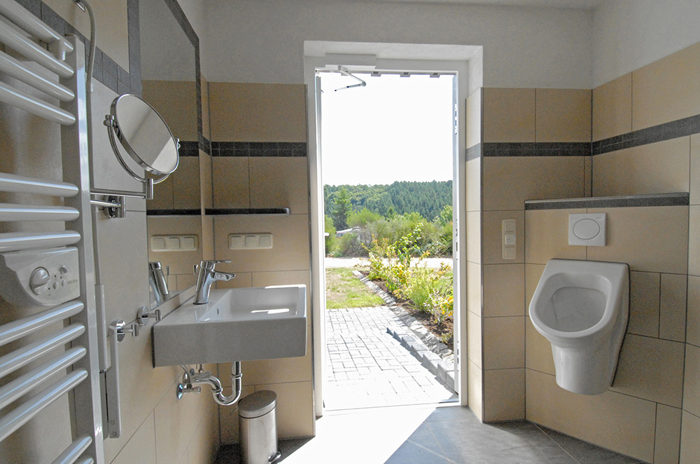 Eine Toilette mit Waschbecken, Urinal, Spiegel, Handtuchwärmer und einer offenen Tür, die nach draußen in einen Gartenbereich bei Tageslicht führt.