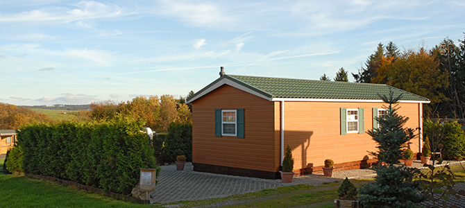 Een kleine modulaire woning van één verdieping met groene luiken en dak staat op een aangelegde kavel met hagen, potplanten en een oprit van grind tegen een achtergrond van bomen en open hemel.