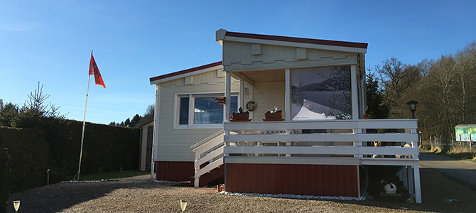 Een klein, crèmekleurig huis met een rode bies, een veranda en een vlaggenmast met een rode vlag, op een grindveld onder een strakblauwe hemel.