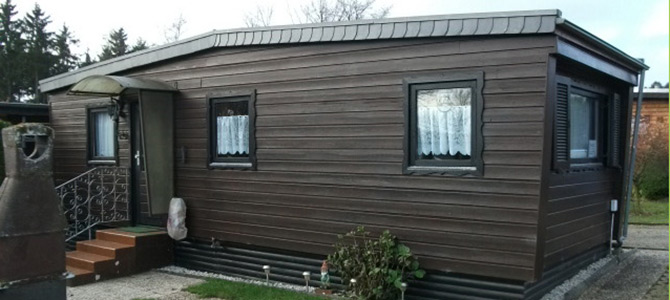Een klein, donkerbruin houten huis met twee ramen aan de voorkant, een zijdeur met trapje en een metalen schoorsteen aan de voorkant.