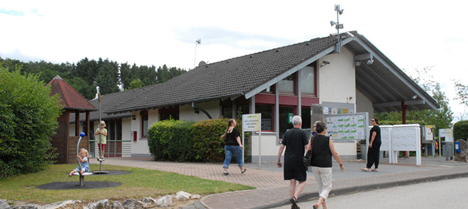 Mensen lopen in de buurt van een modern gebouw van één verdieping met een schuin dak; een kind speelt op een speeltoestel op de voorgrond. Struiken en bomen omringen het gebied.