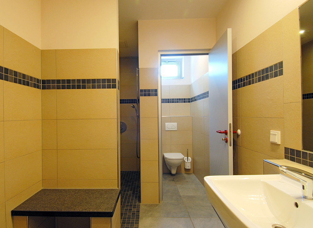 Ein modernes Badezimmer mit beigen Fliesen, einer an der Wand befestigten Toilette, einer Dusche, einem weißen Waschbecken, einem Spiegel und einem kleinen Fenster über der Toilette.