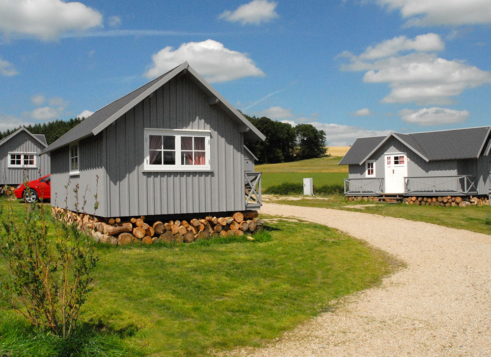 Drei kleine graue Holzhäuschen mit weißer Verkleidung stehen auf einer Wiese, neben einem Kiesweg, unter einem blauen Himmel mit vereinzelten Wolken.