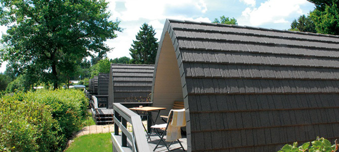 Zwei kleine, graue A-Rahmen-Hütten mit Schindeldächern stehen nebeneinander, umgeben von Grün und Sitzgelegenheiten auf einer Holzterrasse.