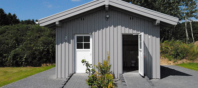 A small, light gray building with vertical siding, two white doors, and a pitched roof, surrounded by greenery and a paved area.