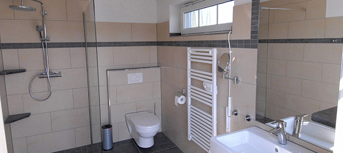 Modern bathroom with a wall-mounted toilet, shower area, towel radiator, sink, and large mirror. Beige tiles with gray accents cover the walls.
