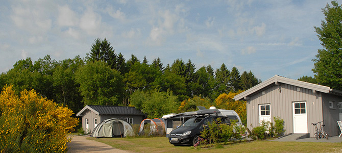 Ein Zeltplatz mit zwei kleinen Hütten, mehreren aufgestellten Zelten, einem geparkten Auto, einem Wohnwagen und einem Fahrrad, umgeben von Bäumen und gelb blühenden Sträuchern unter einem teilweise bewölkten Himmel.