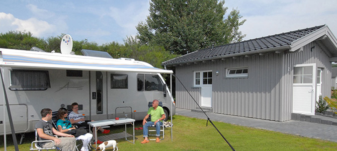 A group of people sit on lawn chairs beside a parked camper van near a small gray cabin, with a dog on the grass in front of them.