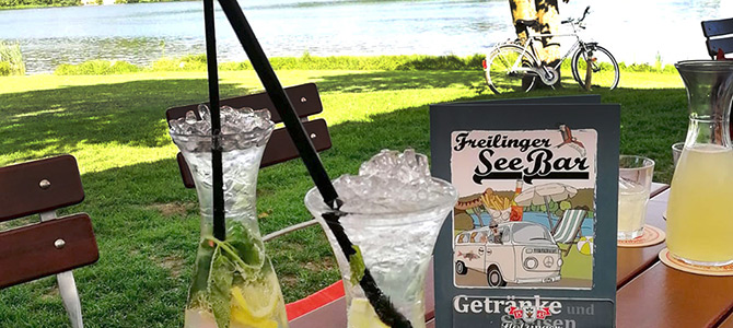 Two glasses of lemonade with ice and mint sit on an outdoor table by a lake, next to a menu labeled Freilinger See Bar, with a bike and water visible in the background.