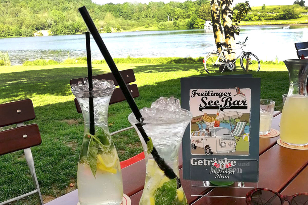 Two glasses of iced drinks with lemon and mint sit on an outdoor table near a lake; a sign reads Freilinger See Bar. Trees, grass, and a bicycle are visible in the background.