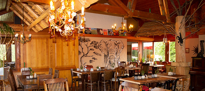 Rustic restaurant interior with wooden beams, eclectic chandeliers, wooden tables and chairs, and a large elephant mural on the wall.
