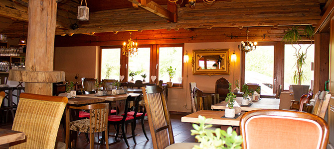Rustic restaurant interior with wooden beams, mixed chairs, decorated tables, large windows, and warm lighting.