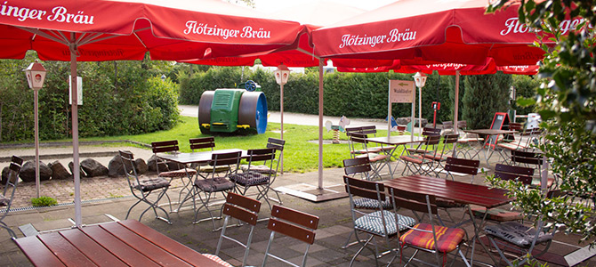 Outdoor seating area with wooden tables and chairs under red umbrellas, surrounded by greenery, with recycling bins visible in the background.
