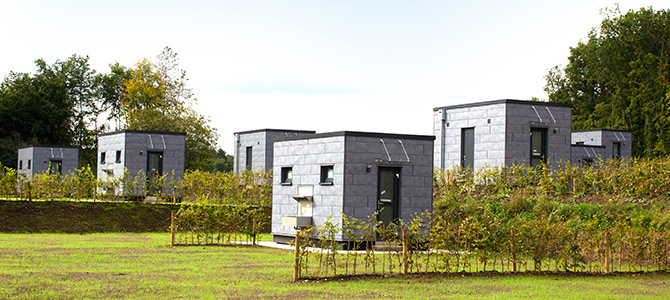Fünf kleine, moderne, graue Modulhäuser stehen im Abstand voneinander auf einem grasbewachsenen Grundstück, das von Bäumen und niedrigen Hecken umgeben ist.