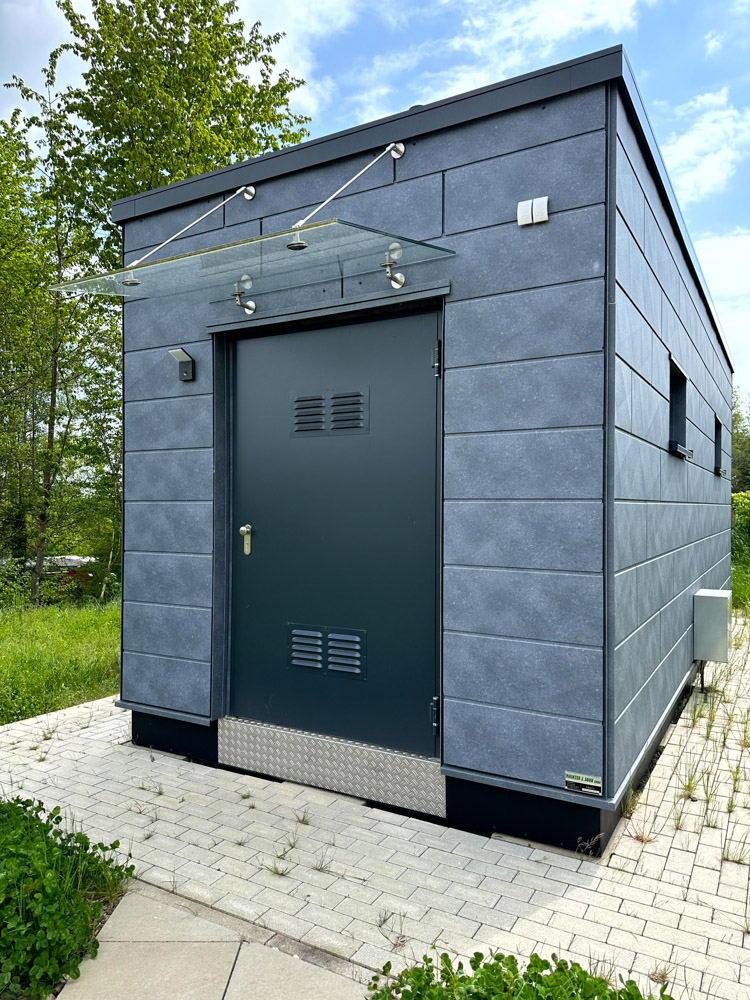 Ein kleines, modernes, graues Versorgungsgebäude mit einer Metalltür, einer Glasmarkise und Lüftungspaneelen auf einer gepflasterten Fläche, umgeben von Gras und Bäumen.
