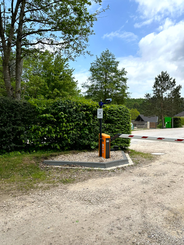 Eine Parkplatzschranke mit einem orangefarbenen Schaltkasten und einer Überwachungskamera steht an einem teilweise bewölkten Tag in der Nähe von Hecken und Bäumen.