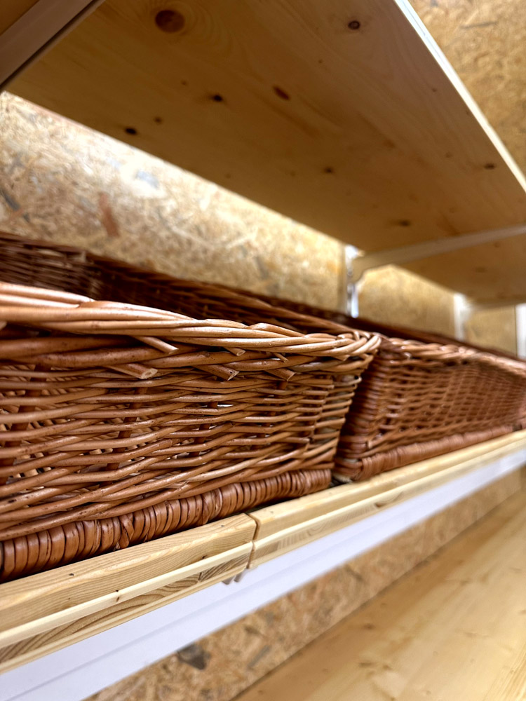 Nahaufnahme von leeren Weidenkörben, die auf Holzregalen in einem Geschäft oder Lagerraum mit holzgetäfeltem Hintergrund angeordnet sind.