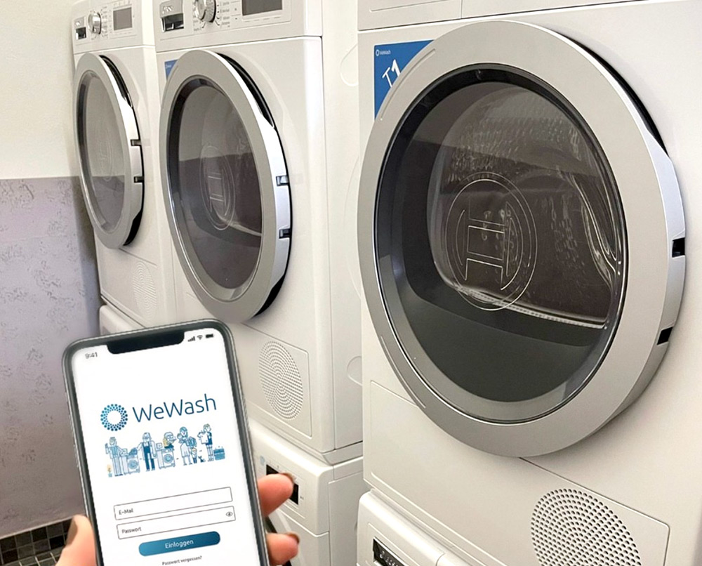 Eine Person hält ein Smartphone mit der WeWash-App vor eine Reihe von gestapelten Waschmaschinen in einem Waschsalon.
