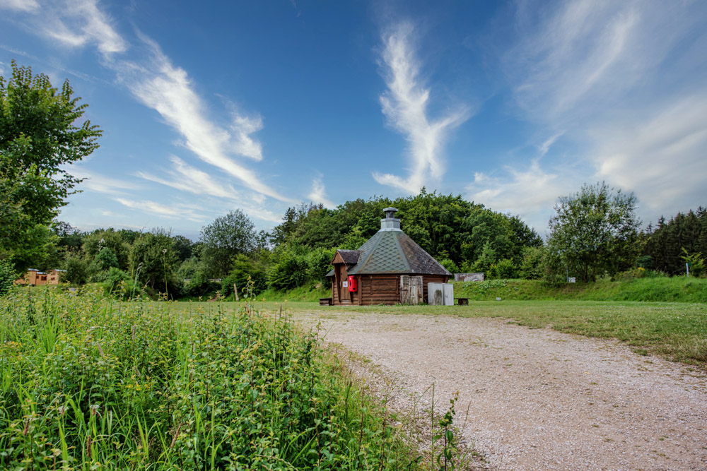 Ein kleines Holzgebäude mit kegelförmigem Dach steht auf einer Wiese neben einem Kiesweg, umgeben von grünen Bäumen und einem blauen Himmel mit Wolkenfetzen.