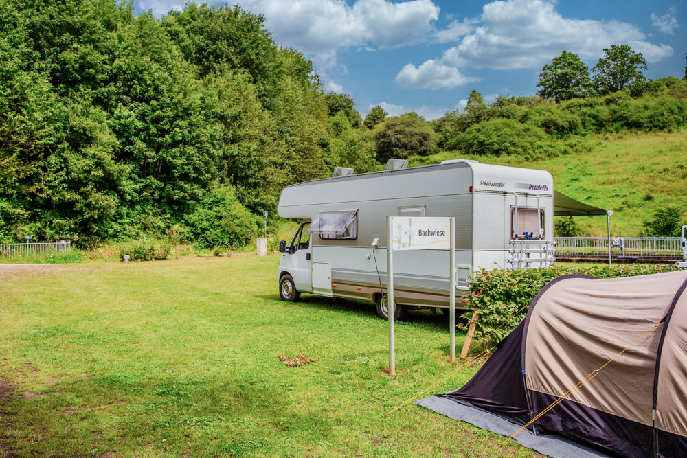Ein weißes Wohnmobil ist auf einem grasbewachsenen Campingplatz in der Nähe von Bäumen geparkt, im Vordergrund stehen ein kleines Zelt und ein Schild mit der Aufschrift Buchen.