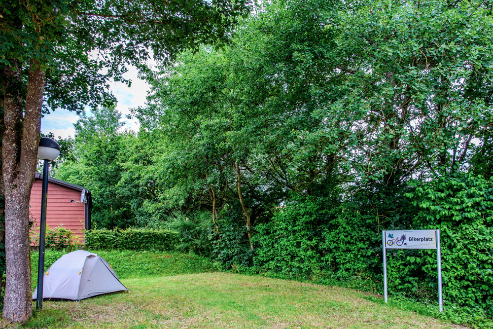 Ein kleines graues Zelt steht auf einer Wiese in der Nähe von Bäumen und einem Gebäude, mit einem Schild mit der Aufschrift Bikeplatz im Hintergrund.
