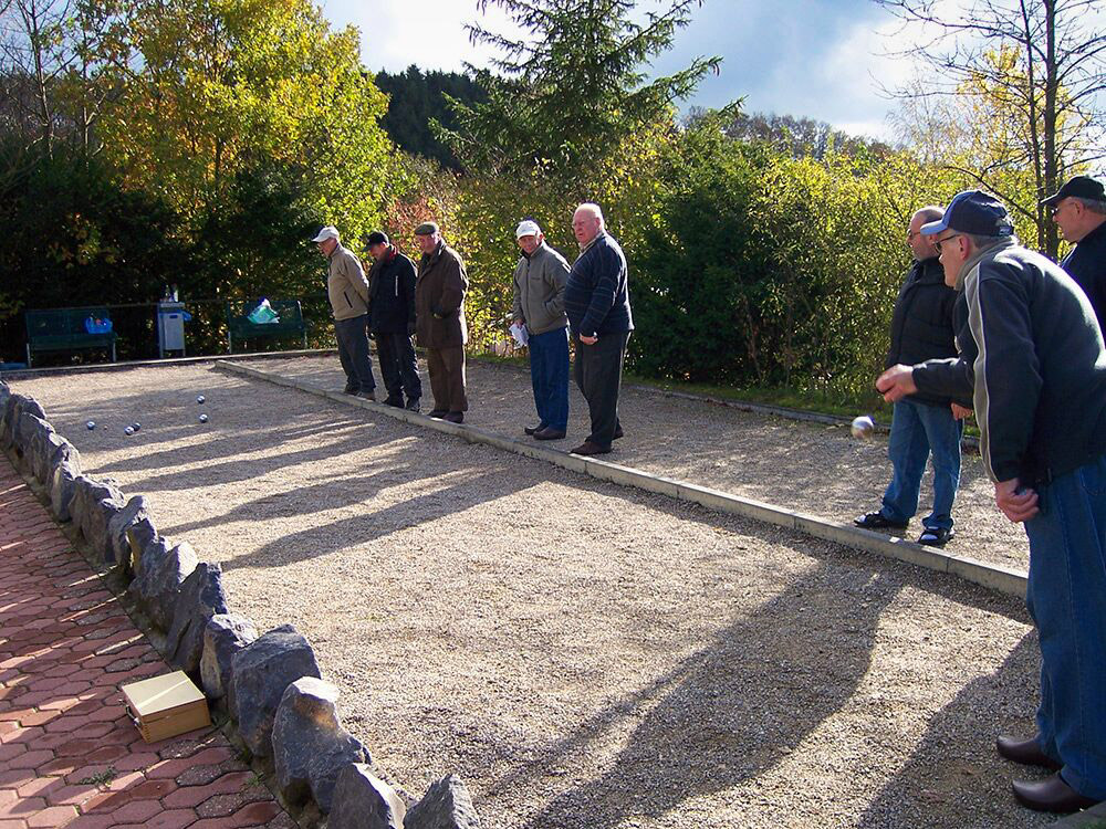 Eine Gruppe von Männern spielt Pétanque auf einem Kiesplatz im Freien, wobei mehrere Metallkugeln auf dem Boden und Bäume im Hintergrund zu sehen sind.