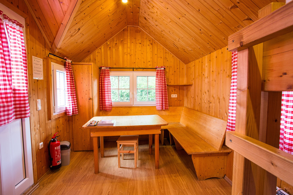 Kleine Holzhütte mit einem Tisch, einer Sitzbank, einem Hocker und rot karierten Vorhängen an den Fenstern; Feuerlöscher an der Tür.
