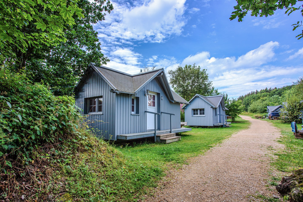 Zwei kleine graue Holzhütten stehen neben einem Kiesweg, umgeben von Grün und Bäumen unter einem teilweise bewölkten blauen Himmel.