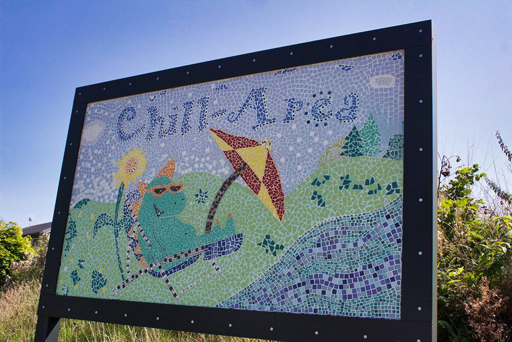 Ein farbenfrohes Mosaikschild mit der Aufschrift "Chill Area" zeigt eine entspannte Figur, die sich in einer sonnigen Graslandschaft unter einem Sonnenschirm räkelt.