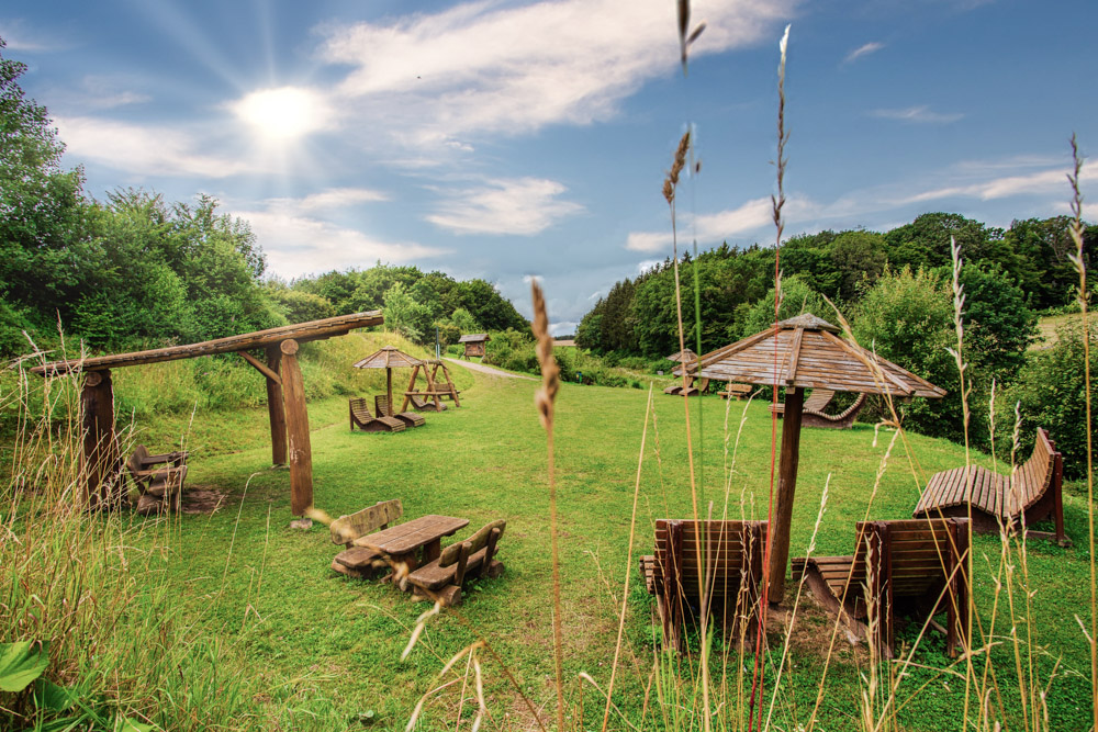 Ein grasbewachsener Außenbereich mit Holzbänken, Liegestühlen, Schaukeln und strohgedeckten Sonnenschirmen, umgeben von Bäumen unter einem hellen, teilweise bewölkten Himmel.