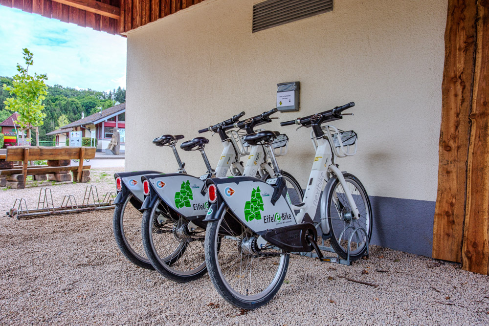 Vier weiße Elektrofahrräder mit der Aufschrift Eifel e-Bike sind an einer Fahrradverleihstation neben einem Gebäude mit Kiesboden abgestellt.