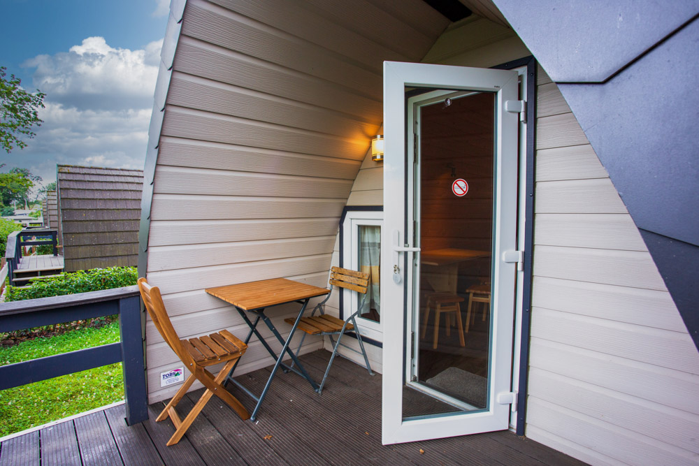 Kleiner Holztisch und zwei Stühle auf einem überdachten Balkon vor einer modernen Kabine mit einer offenen Glastür, die ins Innere führt.