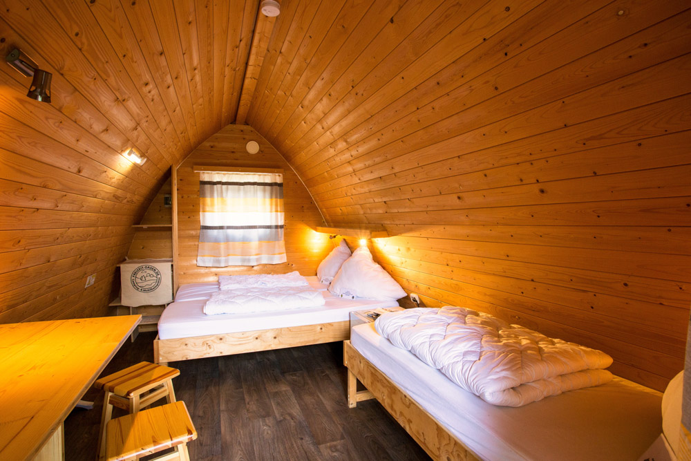 Eine kleine Holzhütte mit schrägen Wänden, die ein Doppelbett, ein Einzelbett, einen Holztisch mit Hockern und ein Fenster mit gestreiften Vorhängen enthält.