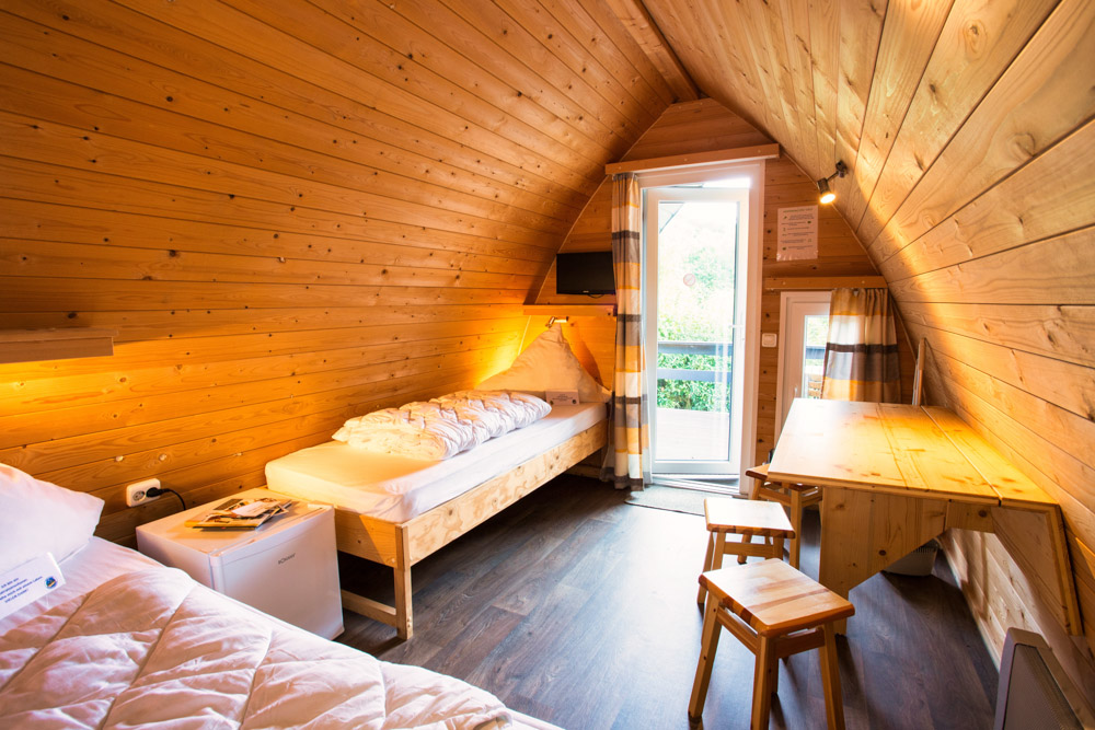 Ein kleines Zimmer im Dachgeschoss mit Holzwänden und -decke, zwei Einzelbetten, einem Schreibtisch mit Hocker, einem kleinen Fernseher und einer Tür, die auf einen Balkon führt, durch den Tageslicht einfällt.